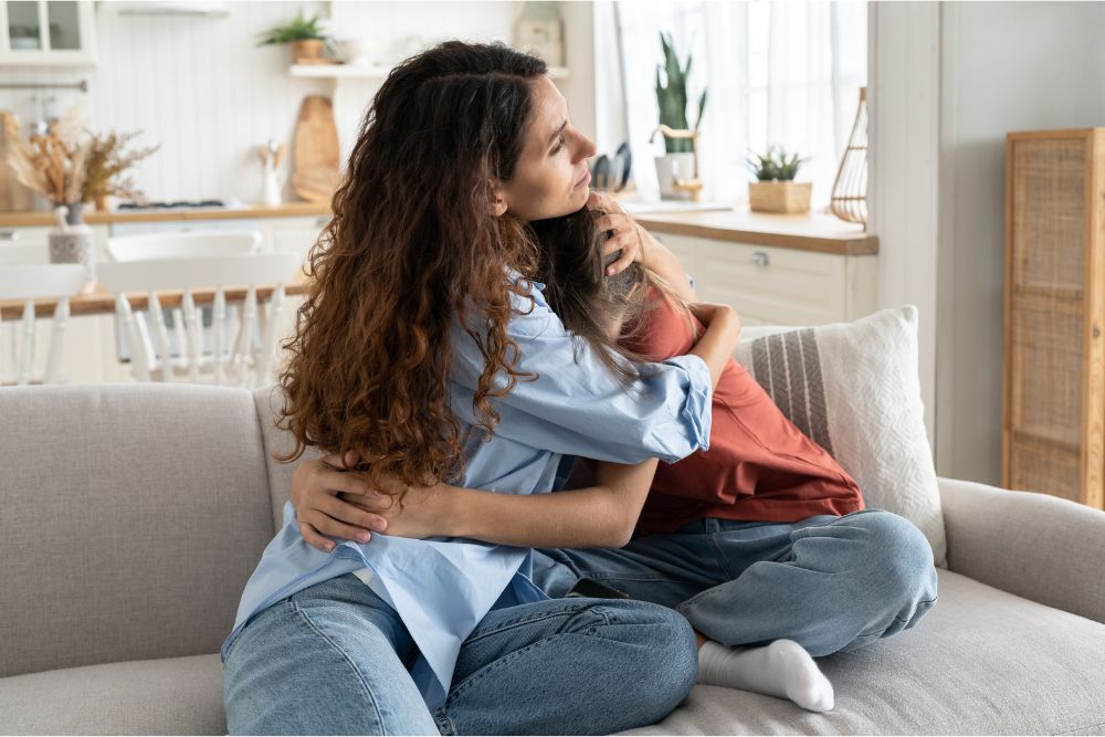 Teen depression treatment often starts with therapy but may require hospitalization. In this image a mother hugs her daughter on a living room couch.