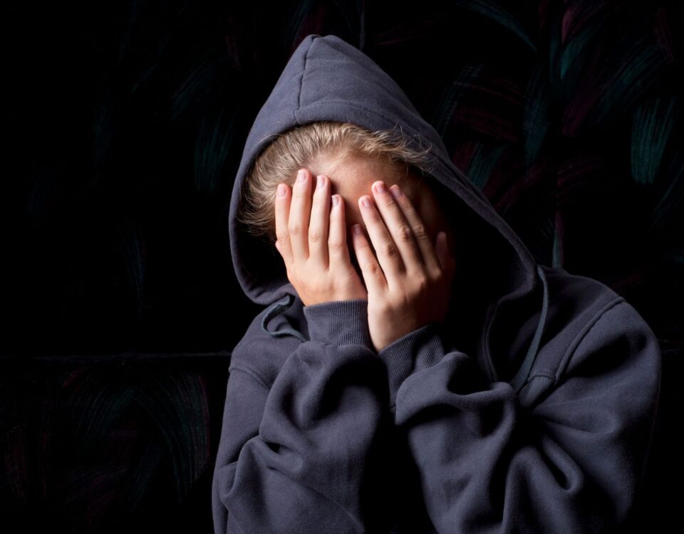 Teenage girl wearing a blue hoodie holds her face in her hands, looking sad and upset.