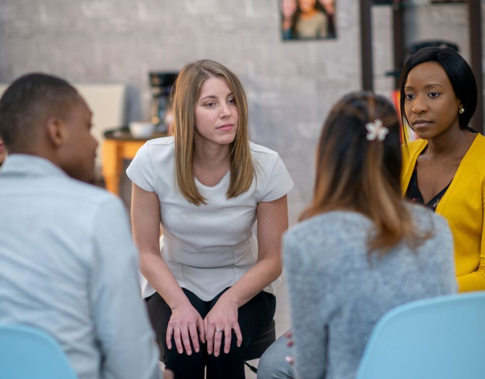 Five young adults sit in chairs forming a circle. They appear to be in a group therapy session.