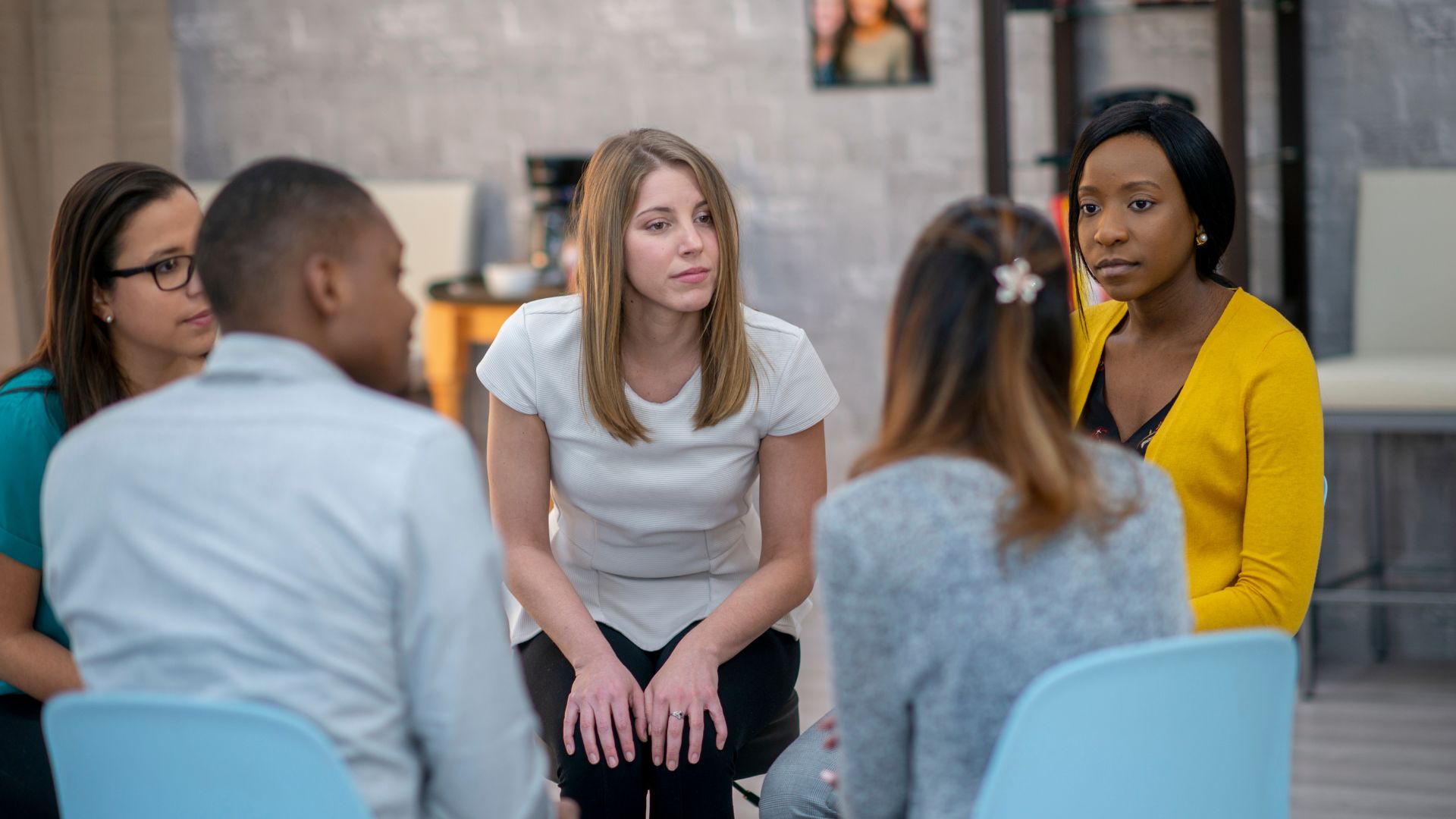 Five young adults sit in chairs forming a circle. They appear to be in a group therapy session.