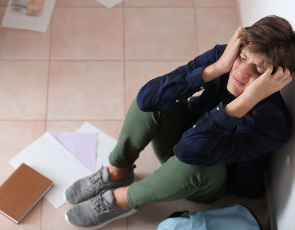 A teenage boy sits on the floor, with a book and papers dropped near him. He is holding his head with both hands, as if he is sad or scared.