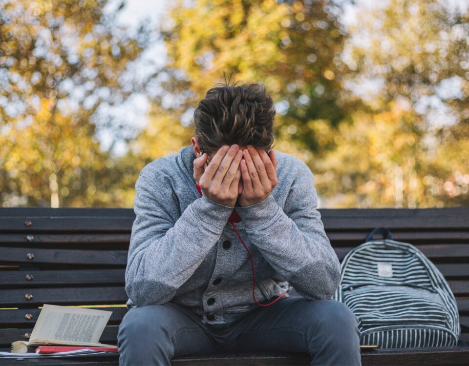 A teenage boy sits on a park bench, covering his eyes with his hands.