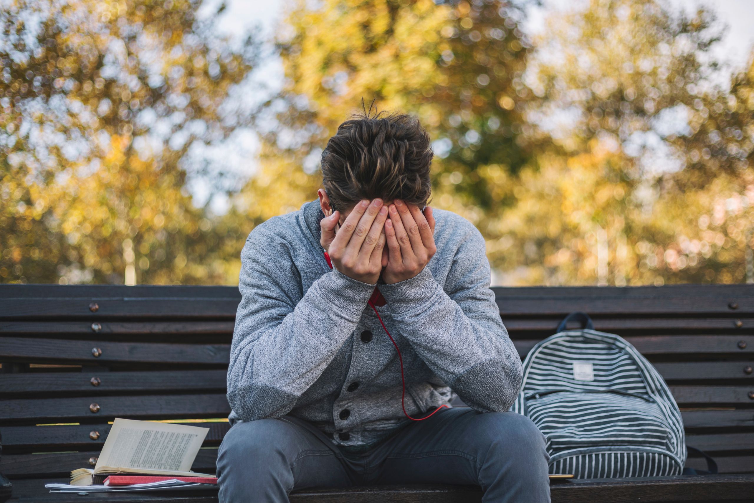 A teenage boy sits on a park bench, covering his eyes with his hands.