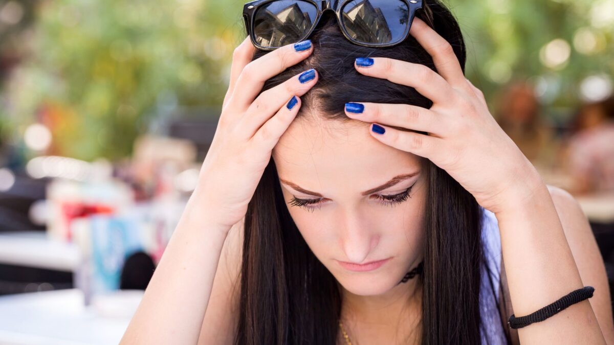 A teenage girl sitting outside holds her hands to the sides of her head, as if she's upset or confused. She is wearing sunglasses.