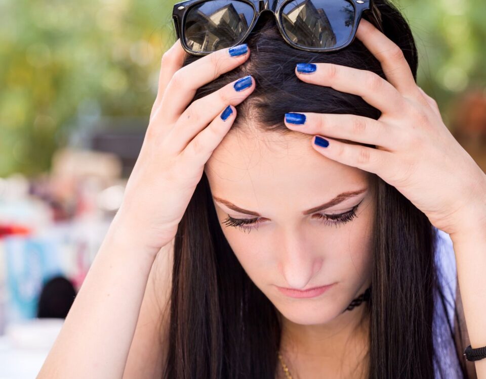 A teenage girl sitting outside holds her hands to the sides of her head, as if she's upset or confused. She is wearing sunglasses.