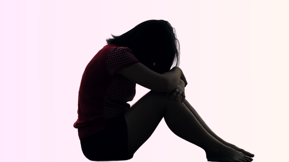 A photo illustration of a girl looking sad, with her head against her knees, sitting in front of a pink background. 