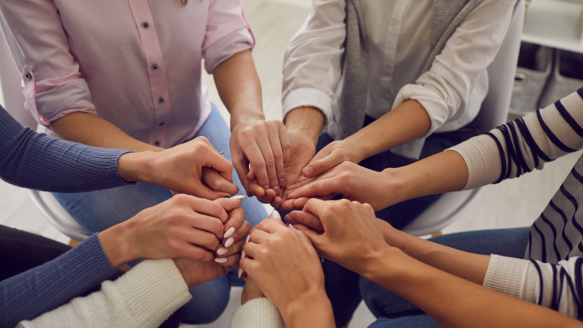 We see a six sets of hands all connecting in a circle of what appears to be six young women in a group therapy session. 