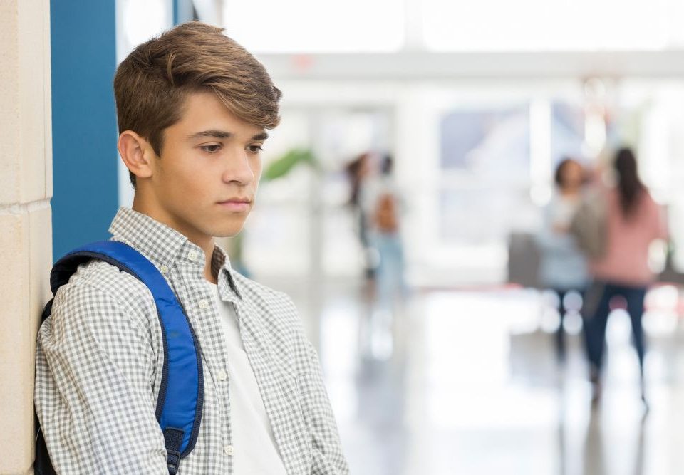 Signs of teen depression include a sense of hopelessness, changes in sleep patterns and personality. In this image a teenage boy leans against a wall in a school hallway looking sad.