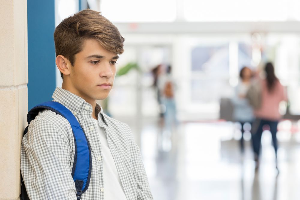 Signs of teen depression include a sense of hopelessness, changes in sleep patterns and personality. In this image a teenage boy leans against a wall in a school hallway looking sad.