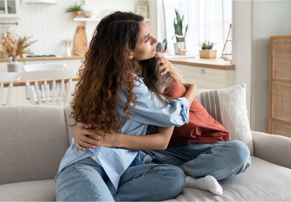 Teen depression treatment often starts with therapy but may require hospitalization. In this image a mother hugs her daughter on a living room couch.