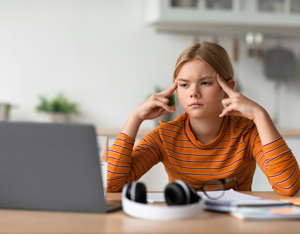 Tired sad concentrated european teen girl presses fingers to head, watches video lesson on laptop