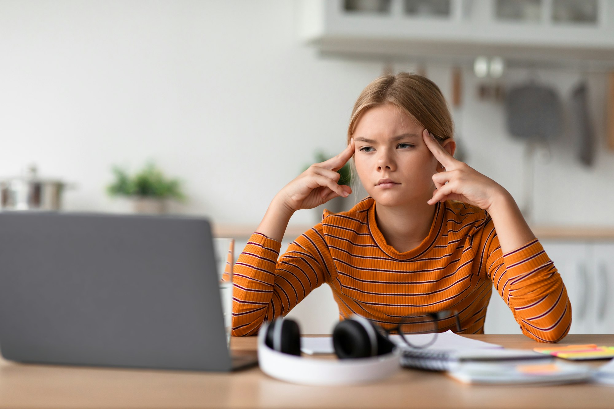 Tired sad concentrated european teen girl presses fingers to head, watches video lesson on laptop