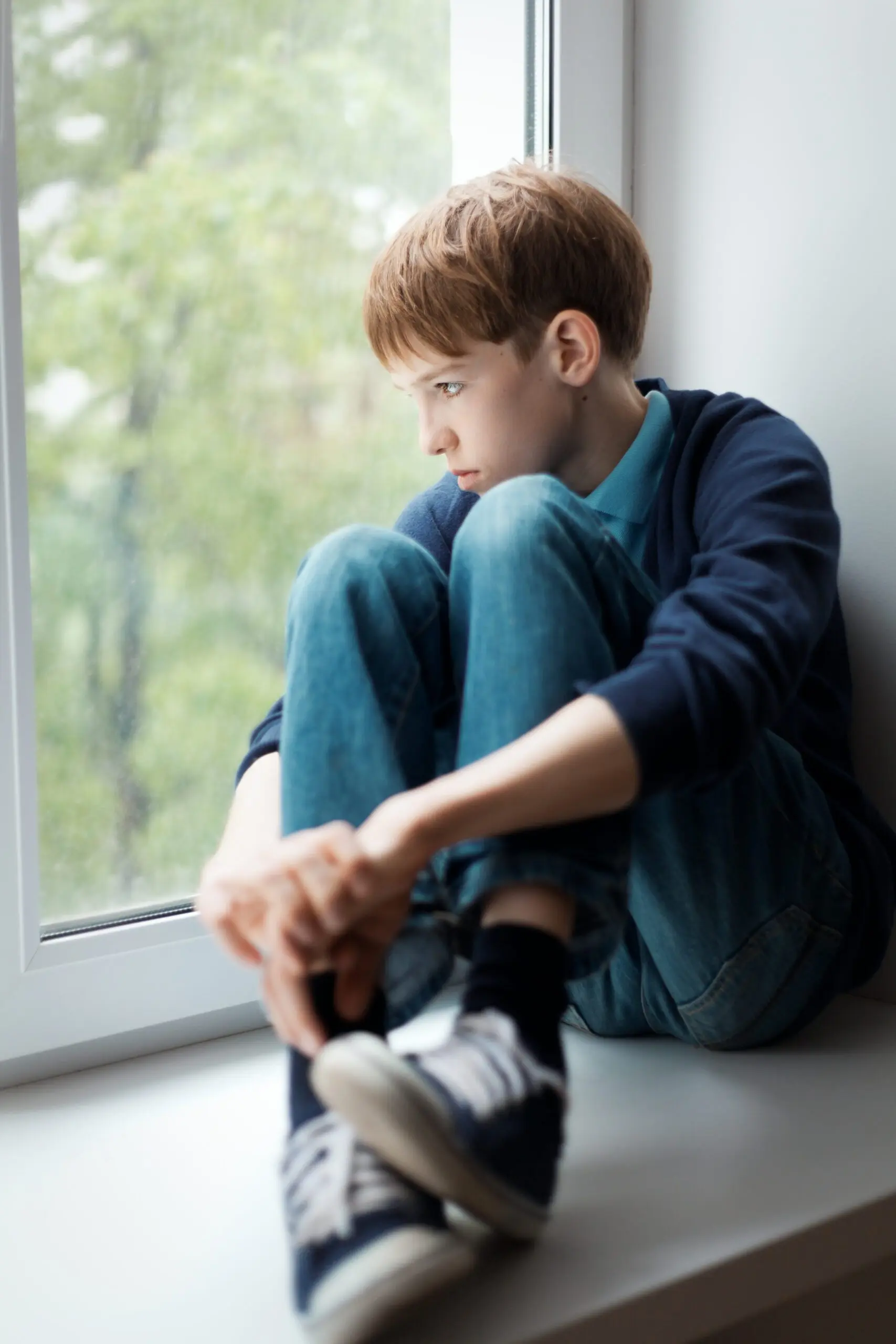 Teen boy sitting in a window with his knees at his chest looking out the window sadly.