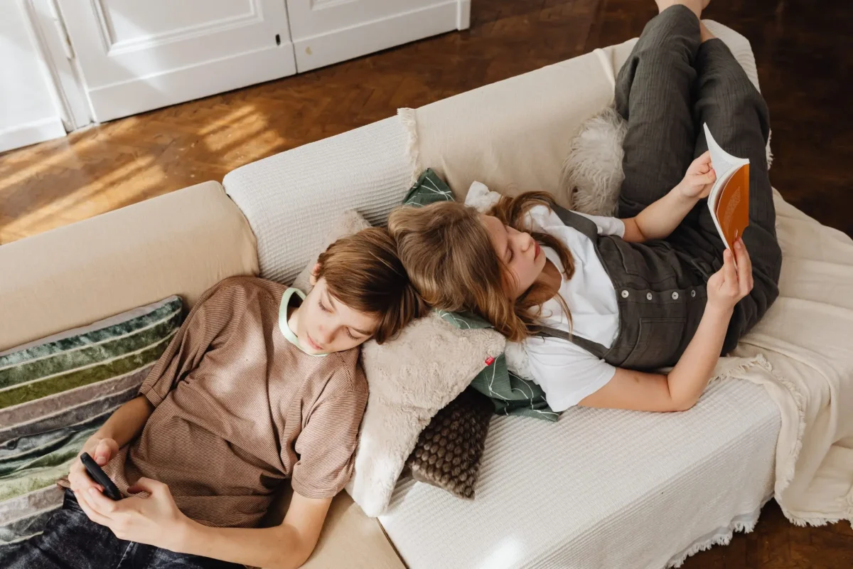 Two siblings reclining on a couch looking at smart phones.