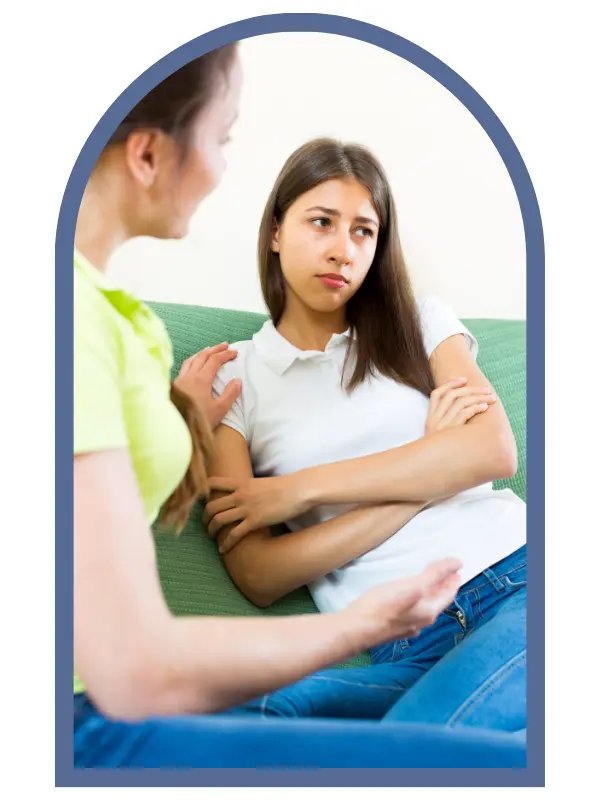 Teen seated with her arms crossed and looking sad. An older person seater beside her, talking.